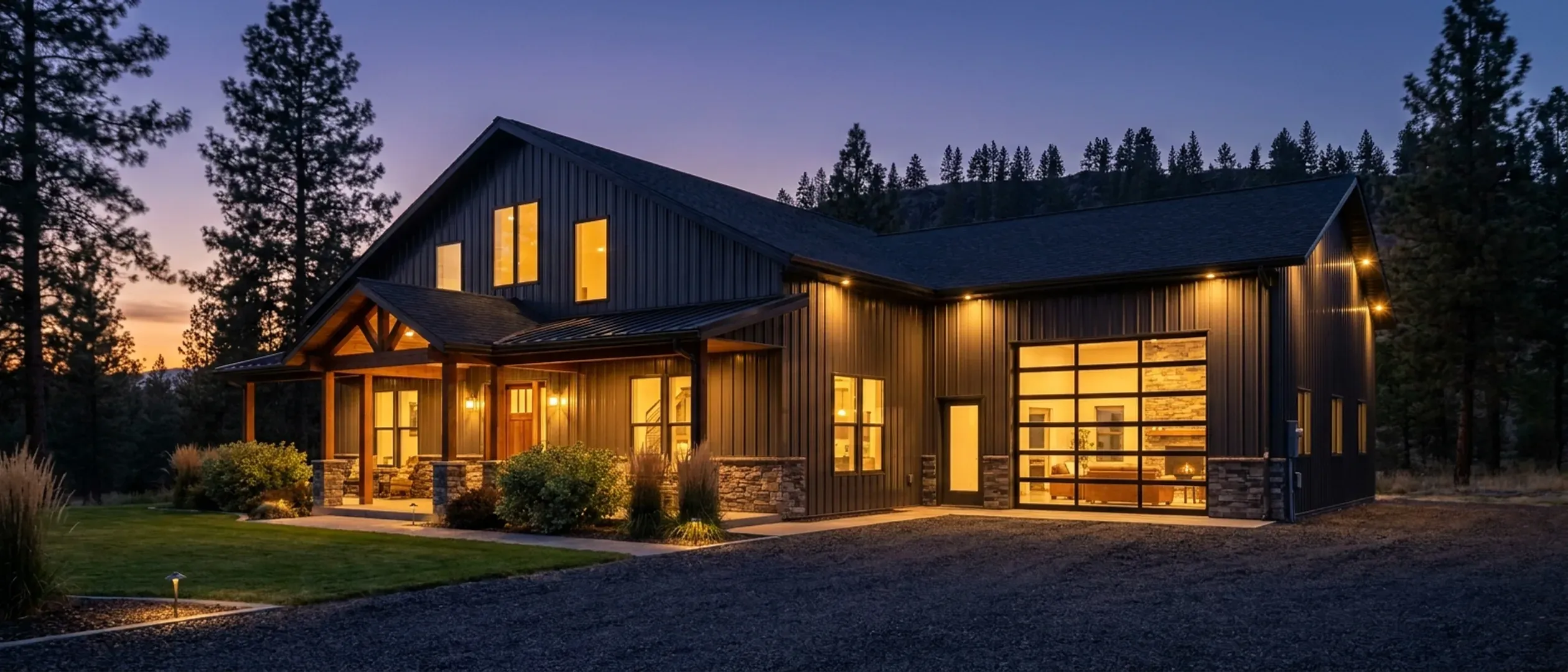 A barndominium at dusk with warm light glowing from every window and exterior lighting along the porch and roofline, tall pines dark against a deep blue twilight sky.
