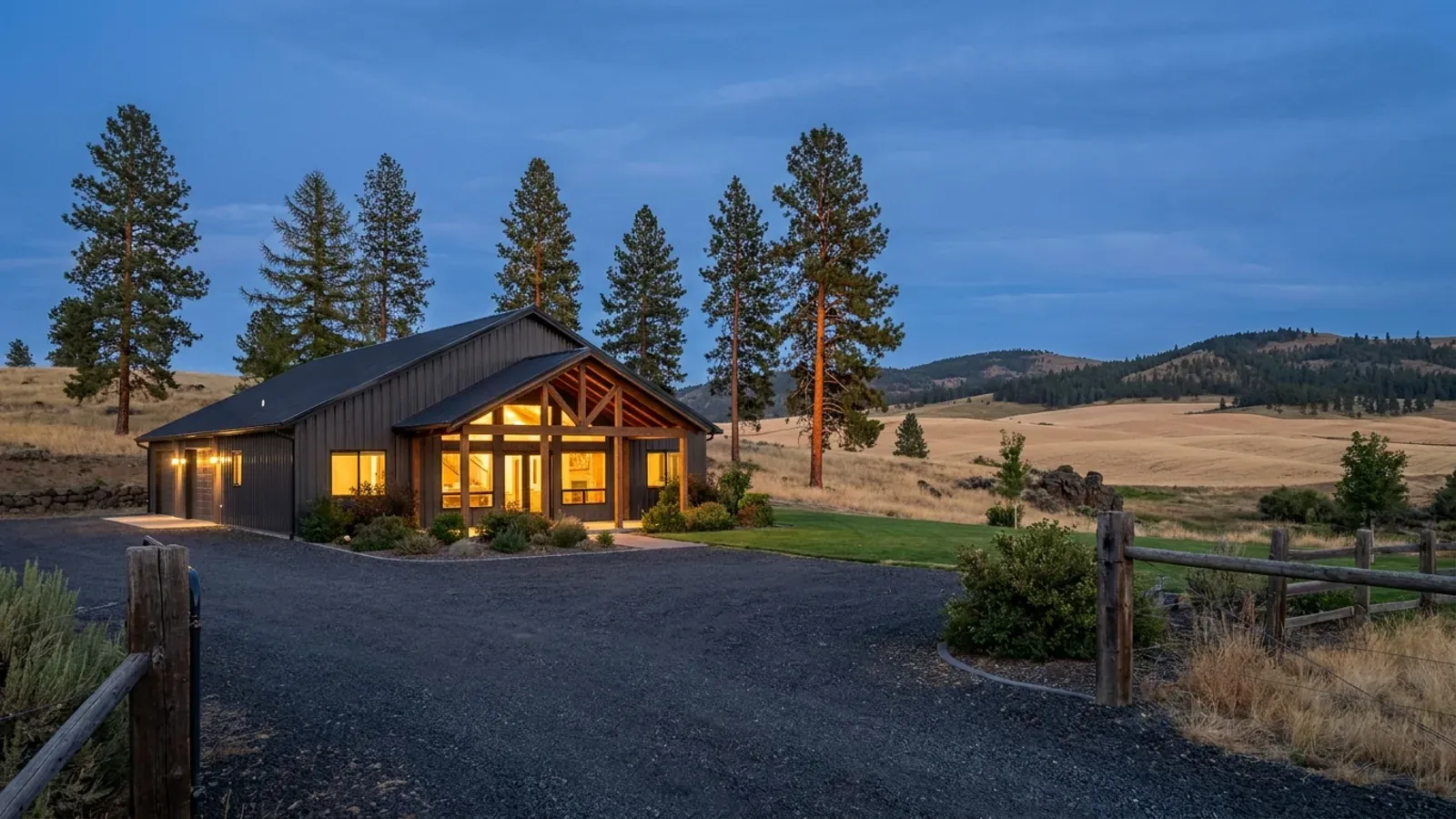 A finished barndominium at dusk with warm interior lighting glowing through its large windows and pines behind.